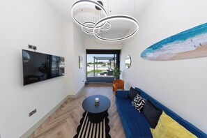A modern living room with a flat-screen TV, a blue sofa, a round coffee table, and a large window overlooking a beach.