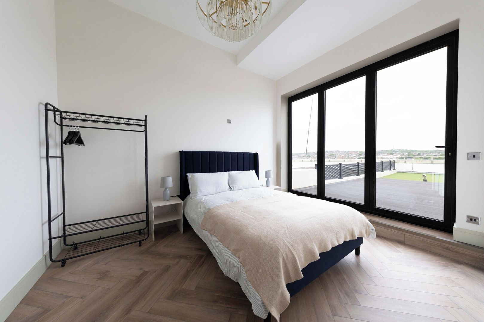 A bedroom with a bed, a nightstand, a clothing rack, and a large window offering a view of the outdoors.