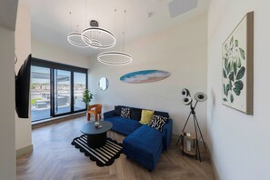 A modern living room with a blue sofa, a round coffee table, and a large window offering a city view. - The Brighton Skyline 2BR Penthouse (Brighton and Hove)