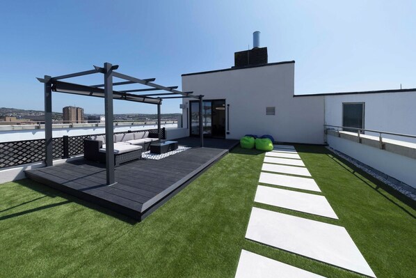 A rooftop terrace with artificial grass, a pergola, outdoor seating, and a view of the cityscape.