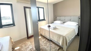 A modern bedroom with a large bed, a bedside table, a mirror, and a window.