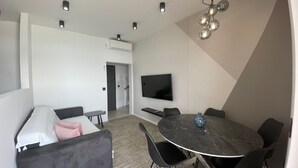 A modern living room with a sofa, a dining table with chairs, a flat-screen TV mounted on the wall, and recessed ceiling lighting.