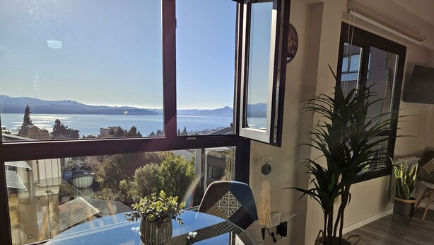 A room with a large window offering a view of a body of water and mountains. There is a glass table with a vase of flowers, a chair, and a potted plant. - Paradise view Bariloche (San Carlos de Bariloche)