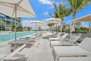 A poolside area with white sun loungers and umbrellas, a clear blue sky, and a modern building in the background.