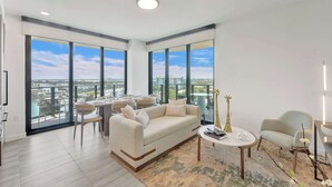 Un salon moderne avec un canapé, une table basse et des fauteuils, de grandes fenêtres offrant une vue sur la ville, et un balcon avec des sièges d’extérieur.
