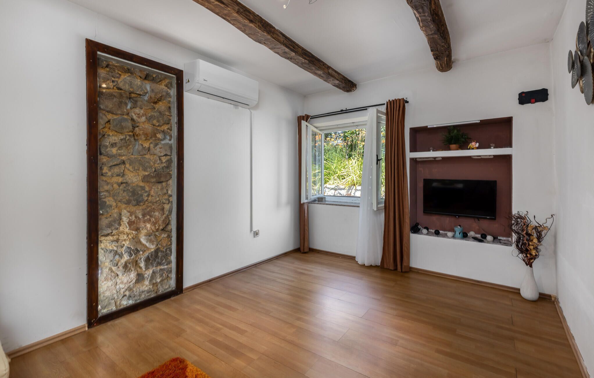 Una habitación con pared de piedra, suelo de madera, televisor montado en la pared y ventana con cortinas.