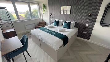 A modern bedroom with a large bed, a desk, and a chair. There is a window with a view of greenery outside.