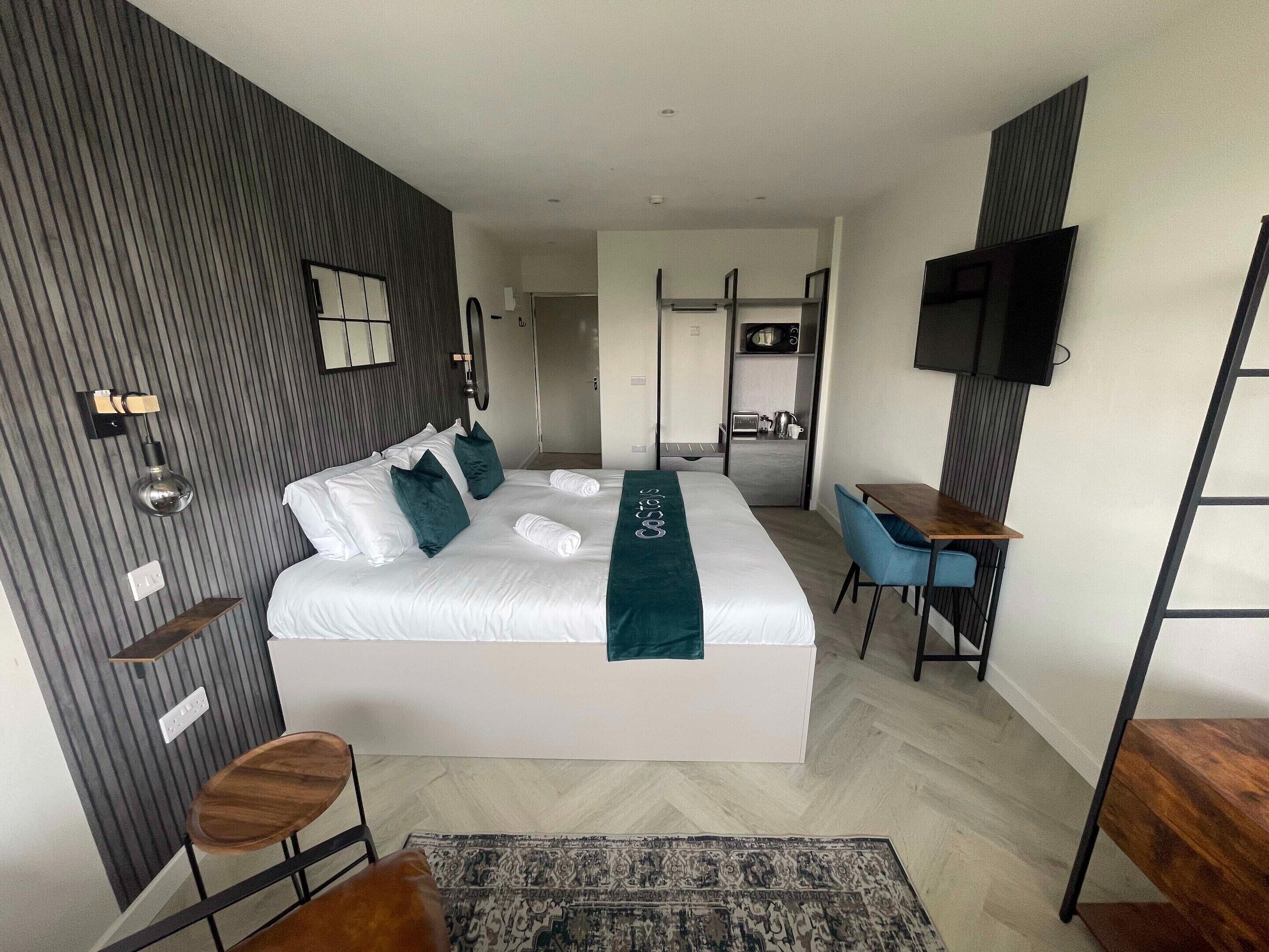 A modern hotel room with a large bed, a desk, a chair, a TV, and a kitchenette.