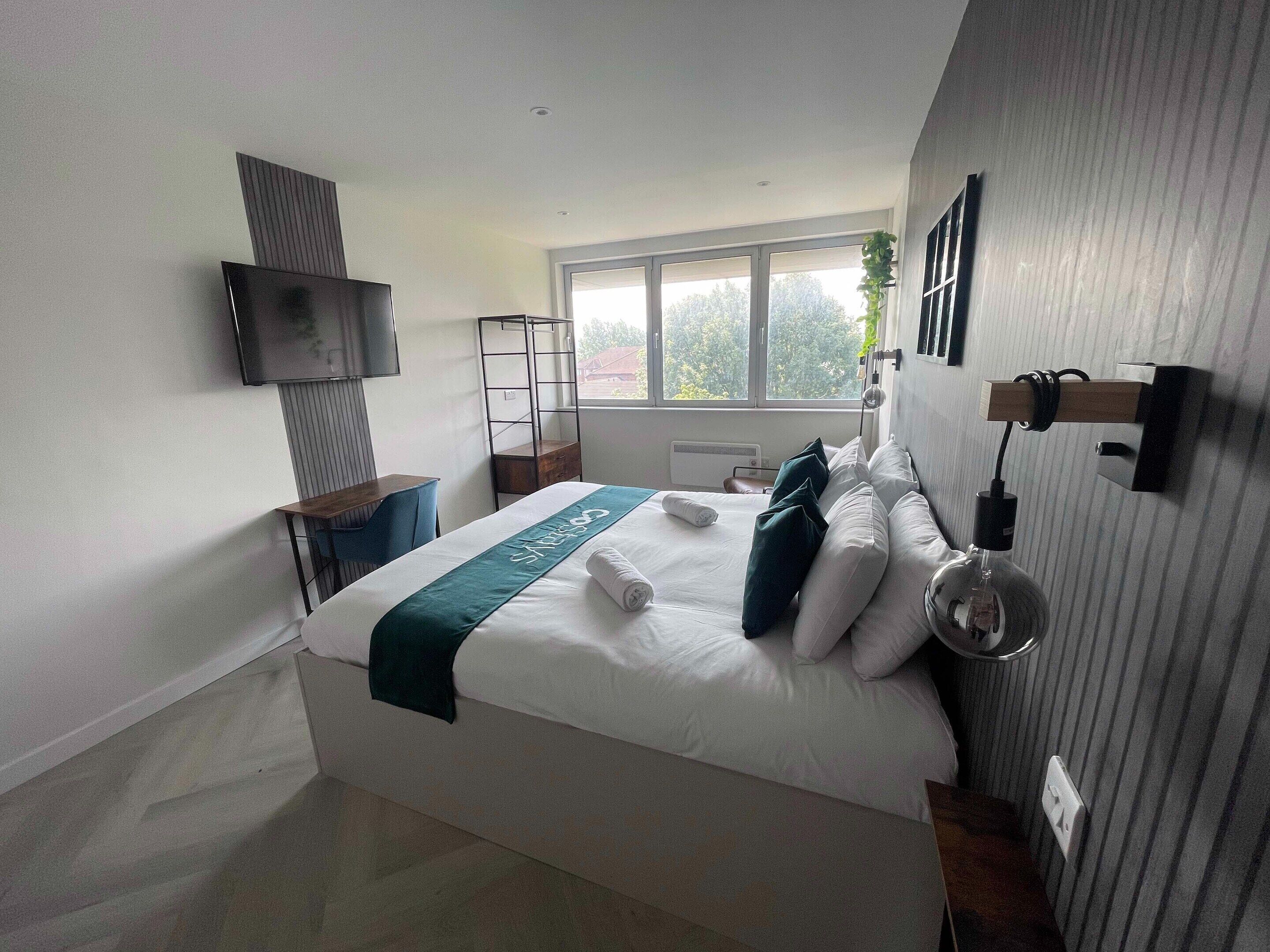 Una habitació d'hotel moderna amb un llit gran, un televisor fixat a la paret i una finestra amb vista a arbres.