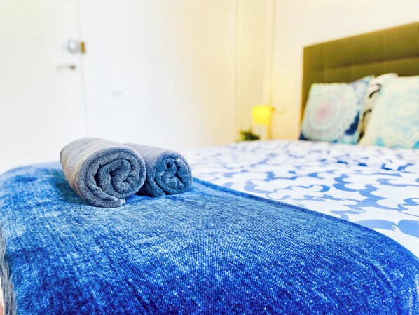 A neatly made bed with a blue and white patterned bedspread and two rolled gray towels on a matching blue pillow.