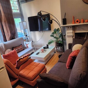 Una sala de estar con sofá, sillón y mesa de centro. Un televisor está montado en la pared, y hay una chimenea con adornos.