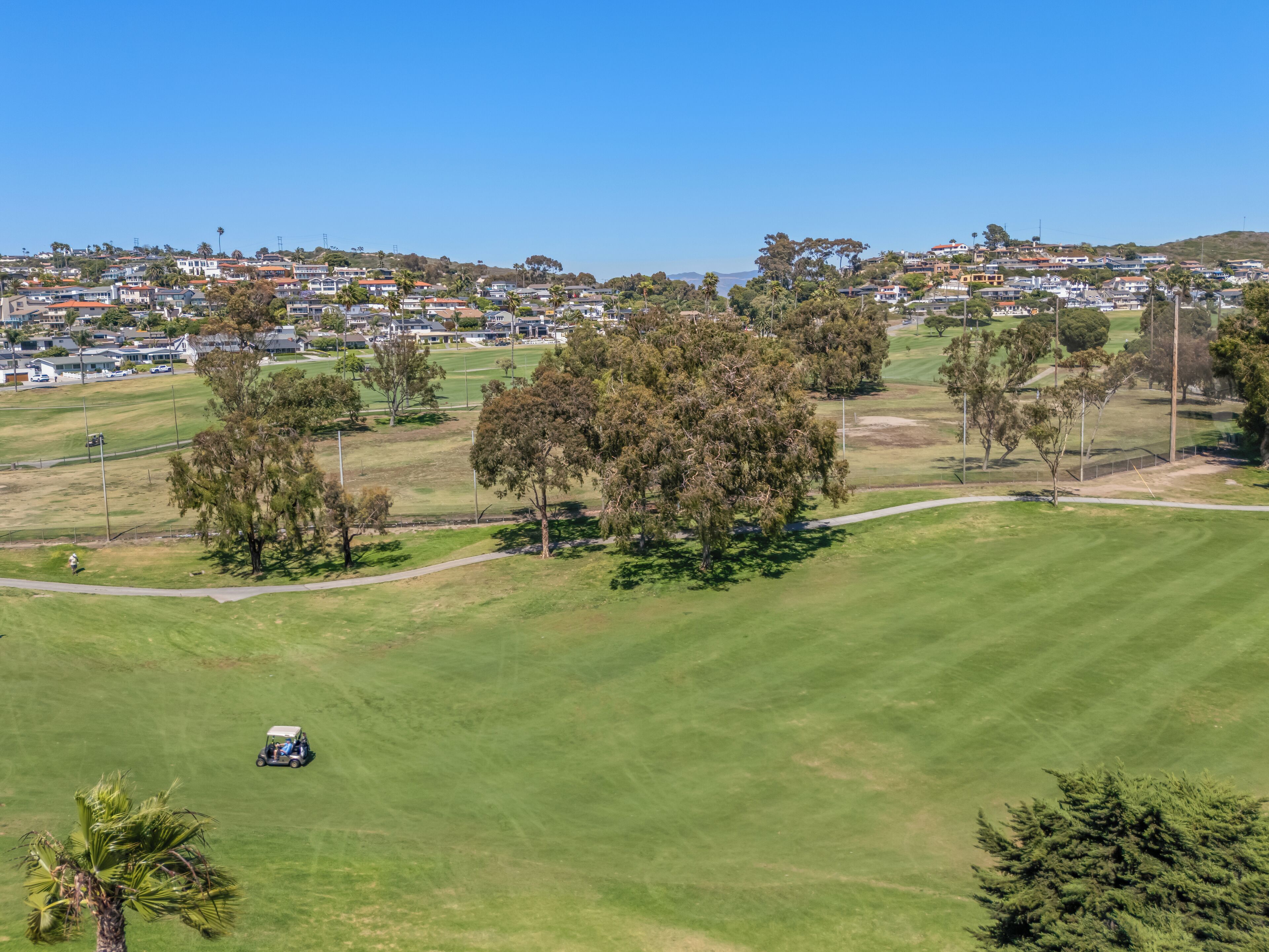 A golf course with a golf cart, surrounded by trees and a residential area in the background.
