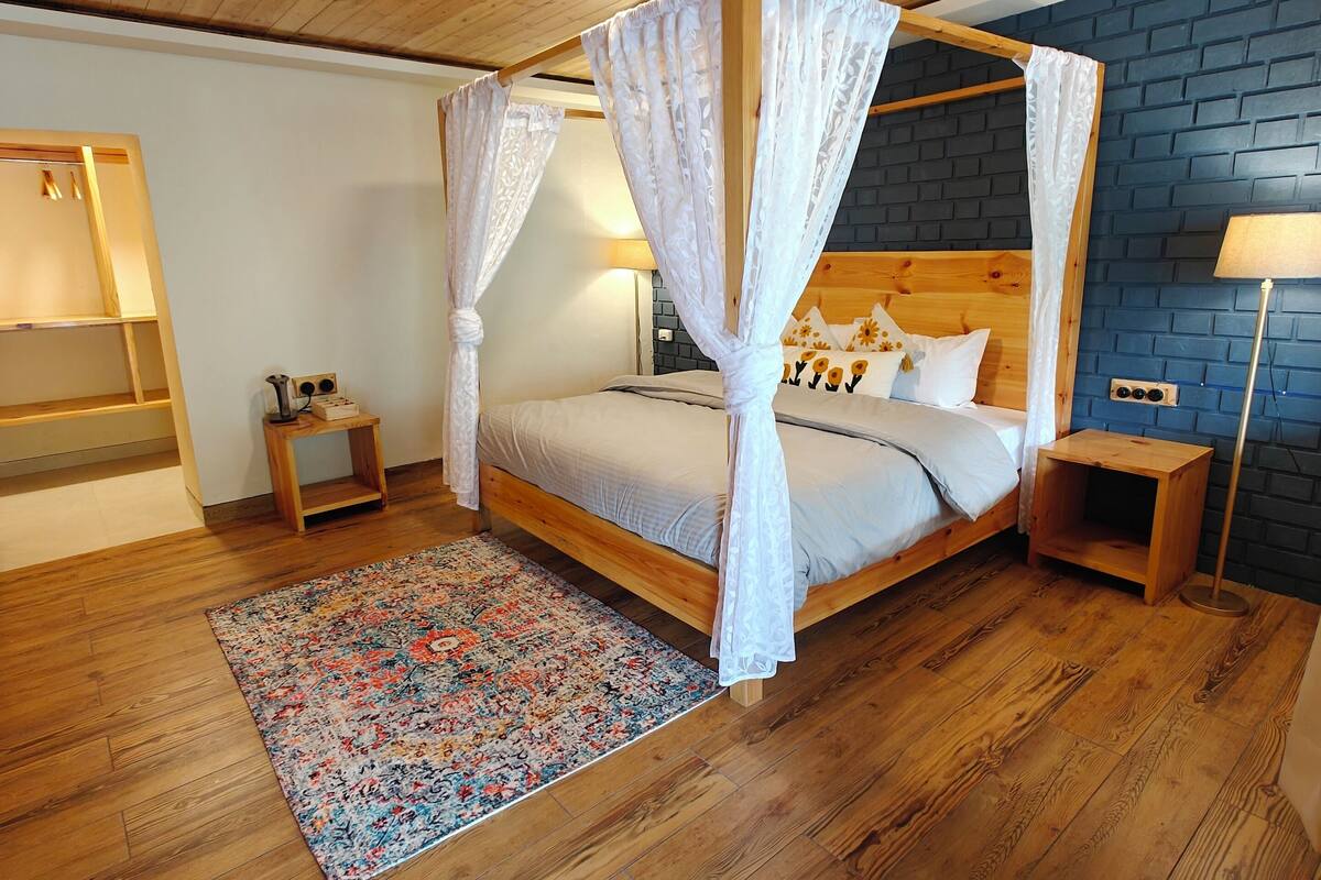 Une pièce avec un plafond en bois, un lit à baldaquin, une petite table de chevet, une commode, une lampe sur pied et un tapis à motifs.
