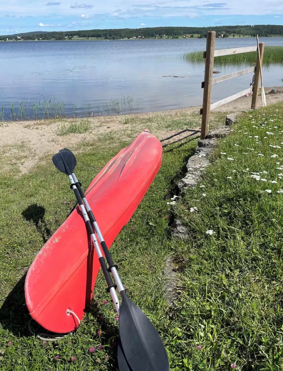 Un kayak rouge, avec une pagaie appuyée contre un poteau en bois, se trouve au bord d’un lac.