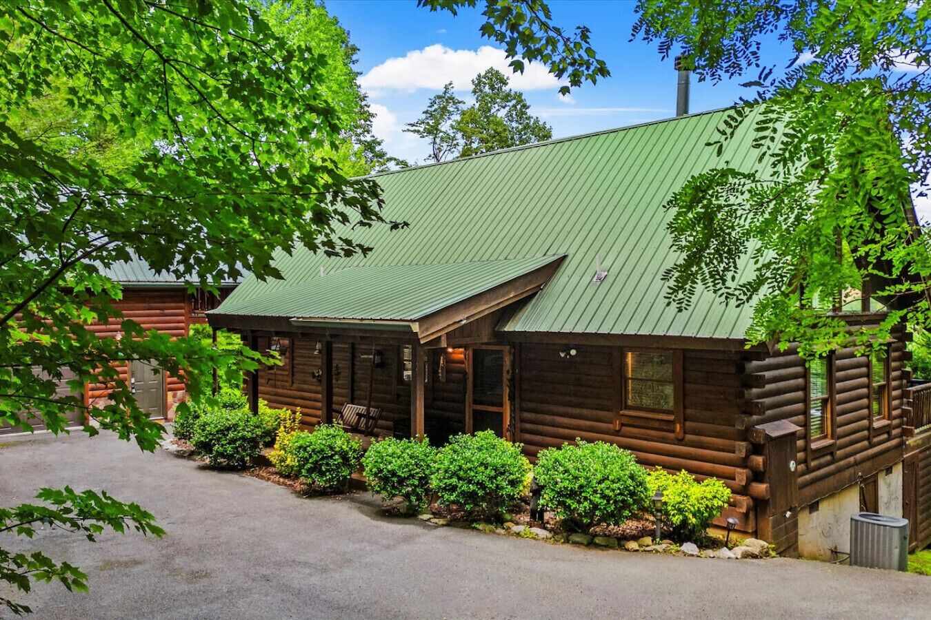 A log cabin with a green metal roof, surrounded by lush greenery and a paved driveway.