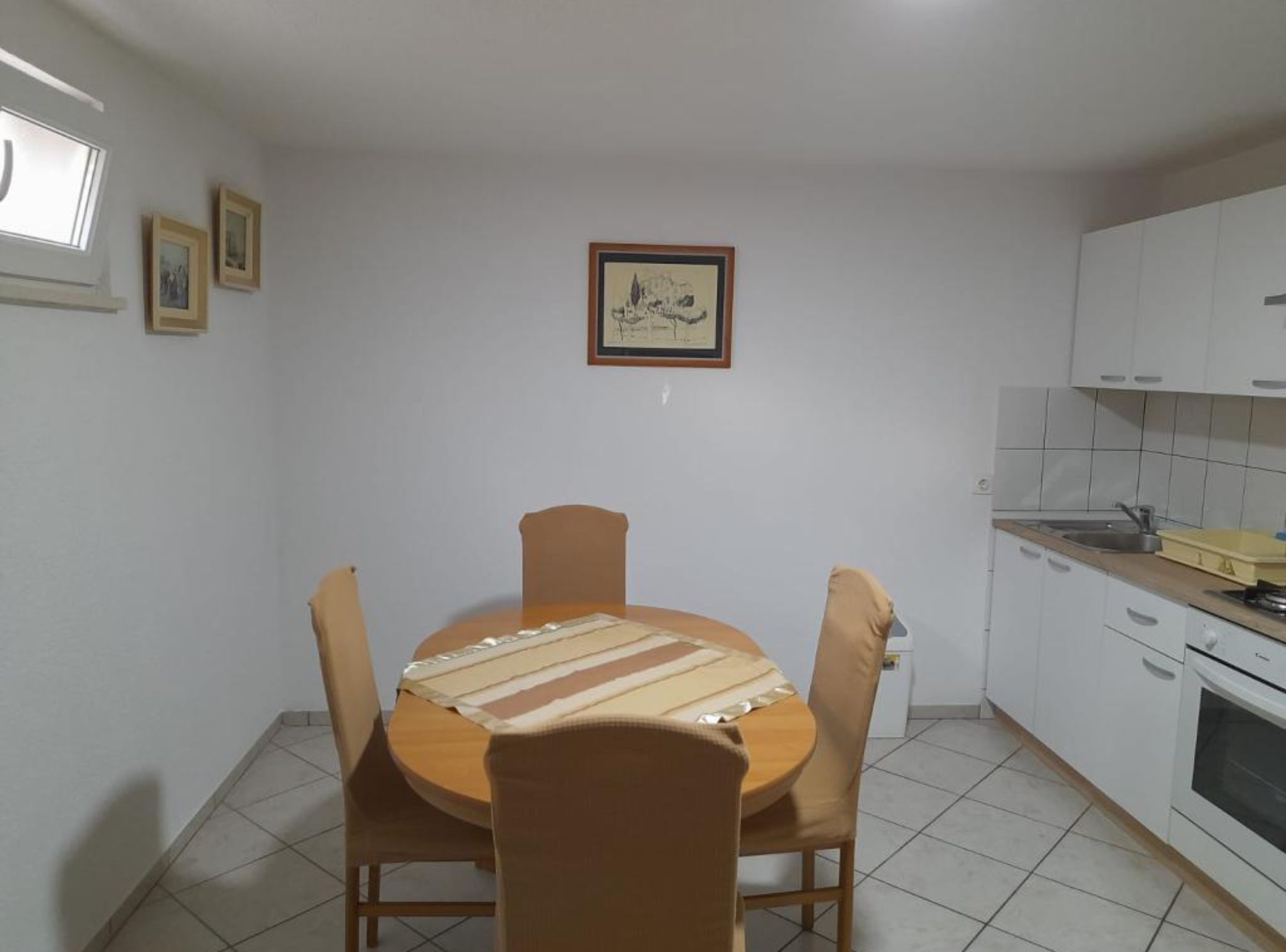 A dining area with a round table, four chairs, and a striped tablecloth. There is a framed picture on the wall above the table.