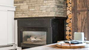 A fireplace with a stone facade, a wooden table with a mug and a book, and a stack of logs against the wall. - Ski-In/Out Luxury•1 Blk to Main•Pool+Hot Tub•Sauna (Breckenridge)