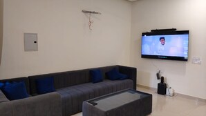 Een moderne woonkamer met een hoekbank, een salontafel met glazen blad en een aan de muur bevestigde televisie.