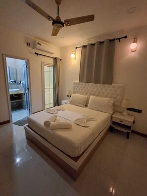 Une chambre à coucher avec un lit, des tables de chevet, un ventilateur de plafond et une salle de bain visible à travers une porte ouverte.