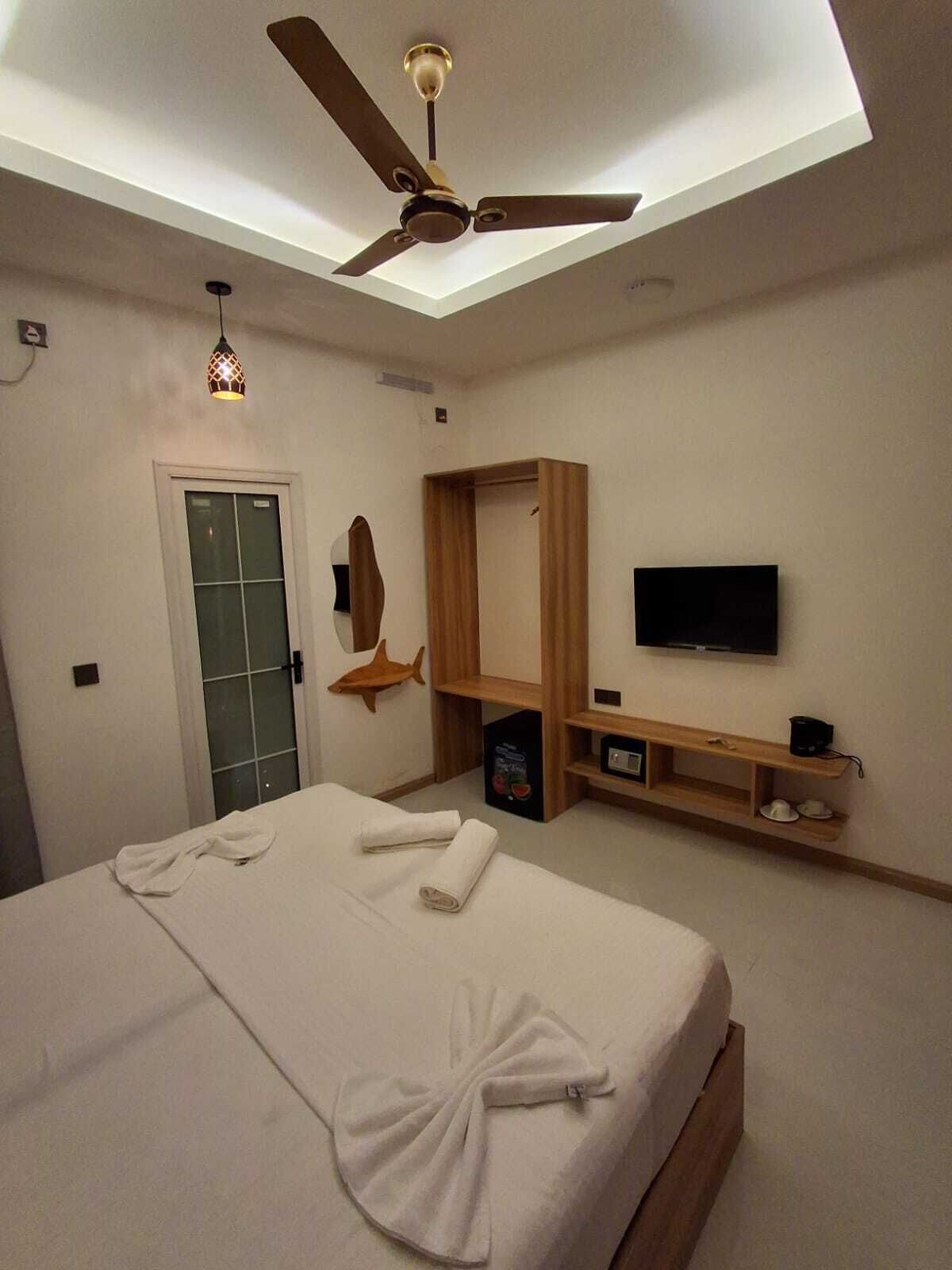 A bedroom with a bed, a ceiling fan, a TV, and a wooden shelf.