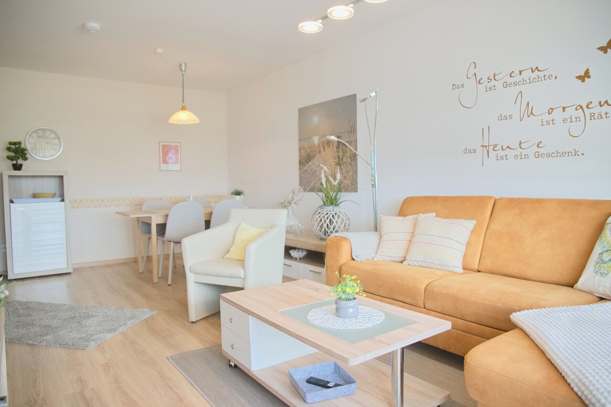 Una sala moderna con un sofá, una mesa de centro, una zona de comedor con una mesa y sillas, y una pared con una cita decorativa.