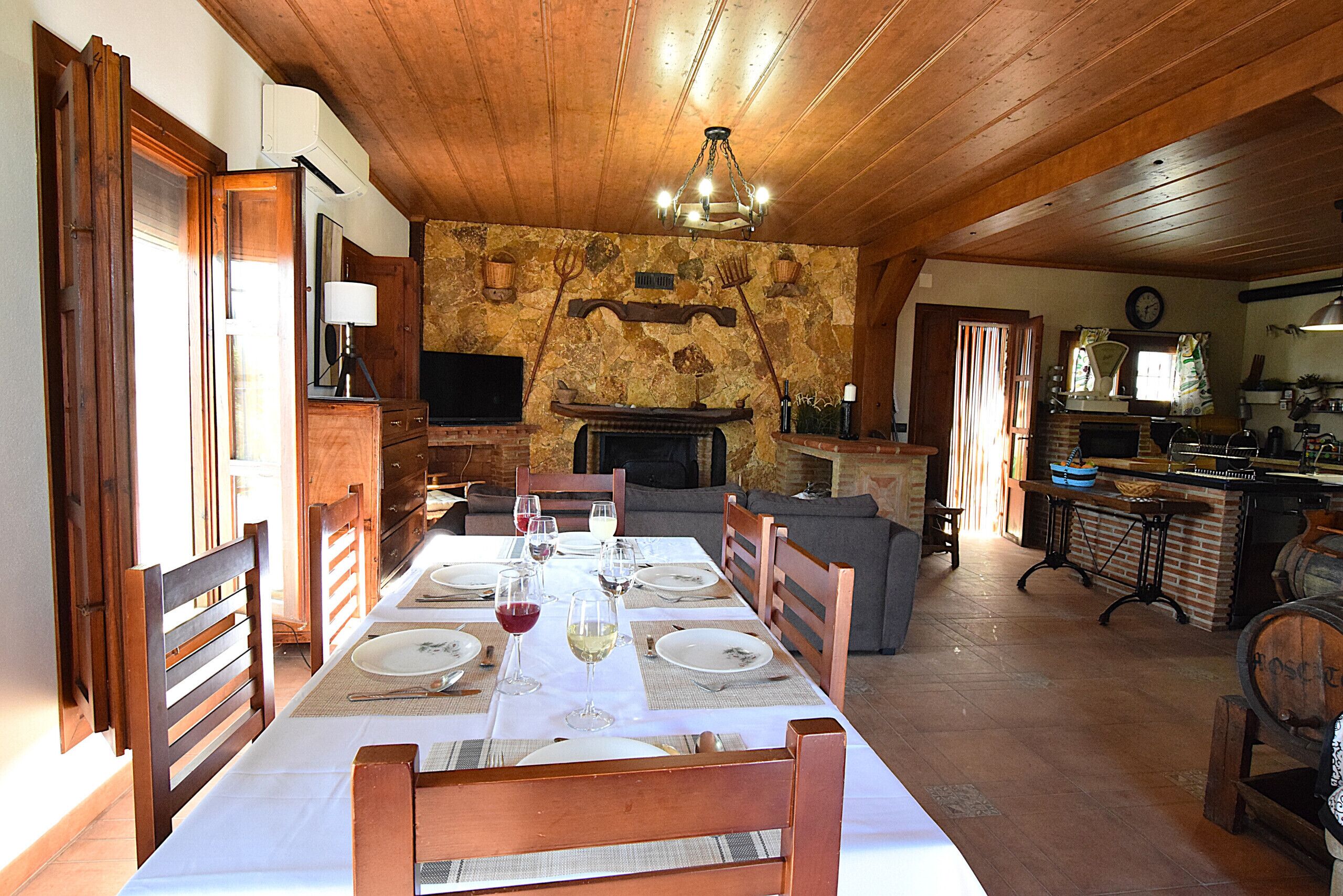 Un comedor con una larga mesa preparada para una comida, una chimenea y una cocina al fondo.