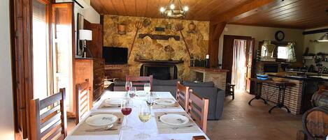 Un comedor con una larga mesa preparada para una comida, una chimenea y una cocina al fondo.