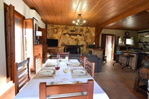 A dining area with a long table set for a meal, a fireplace, and a kitchen in the background. - Idyllic Andalusian villa with private pool & stunning views (Riogordo)