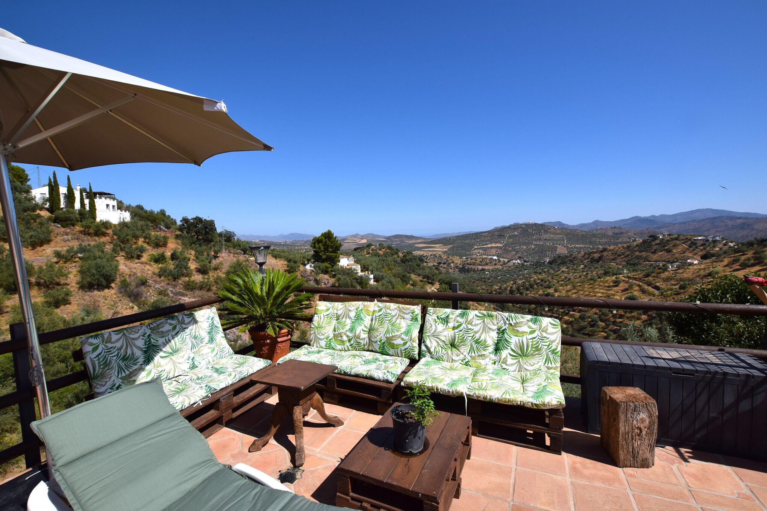 A terrace with a view of a hilly landscape, a white building, and a table with a plant.