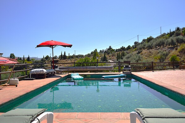 A swimming pool with lounge chairs and a red umbrella, surrounded by a wooden fence and a hilly landscape.