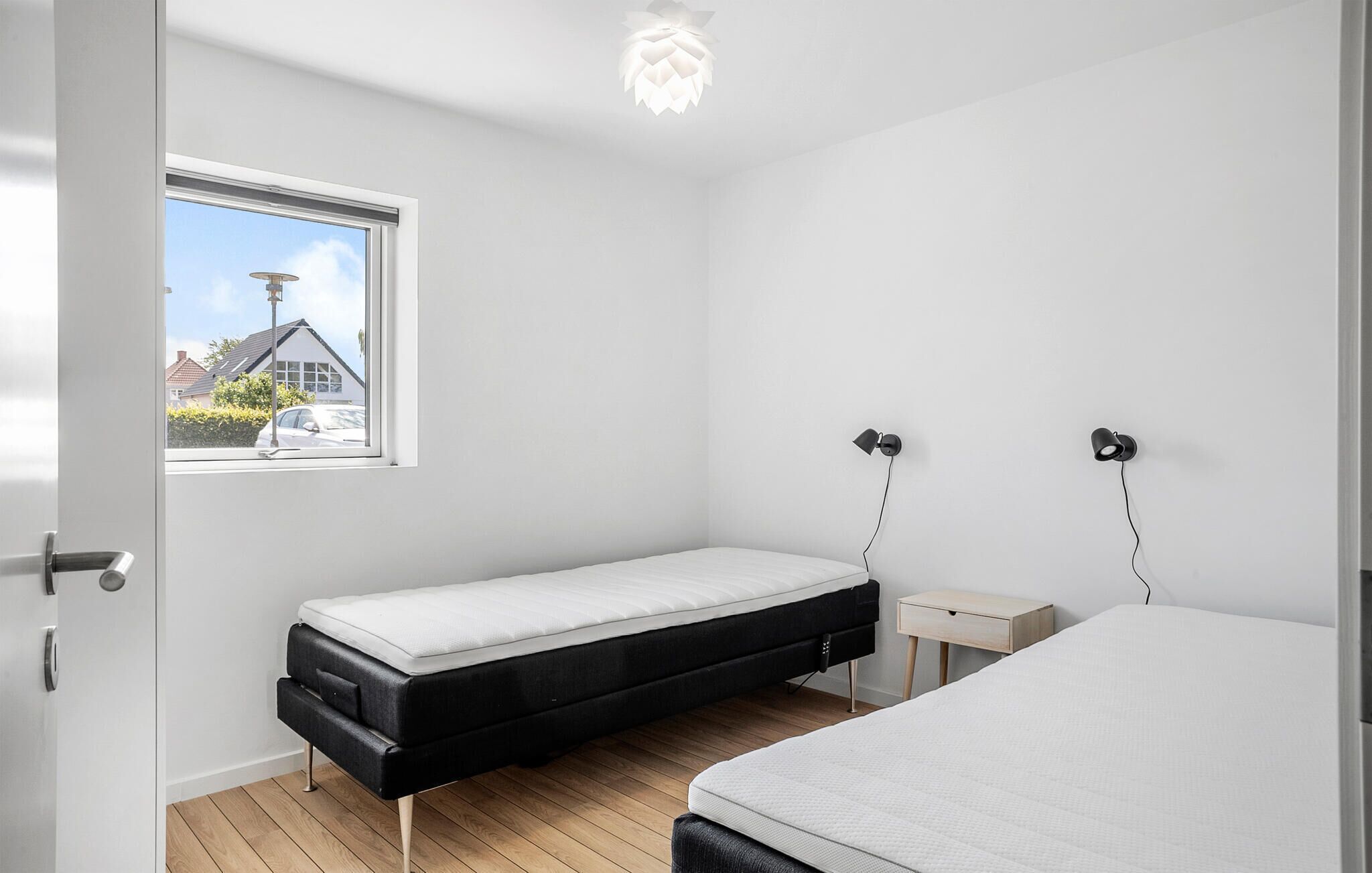 Et soveværelse med to senge, et vindue med udsigt til et hus og et natbord med en lampe.