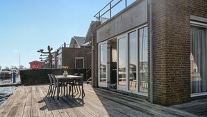 An outdoor patio with a table and chairs, a brick building with large windows, and a view of the water and boats in the background. - Westergeest beneden 2 slaapk (Uitgeest)