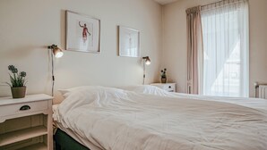 A bedroom with a large bed, two bedside tables with lamps, a potted plant, and two framed pictures on the wall. - Westergeest beneden 2 slaapk (Uitgeest)