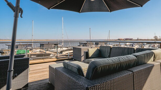 Eine Terrasse mit Sitzgelegenheiten und einem großen Sonnenschirm mit Blick auf einen Jachthafen, wo Boote und eine entfernte Windmühle zu sehen sind.