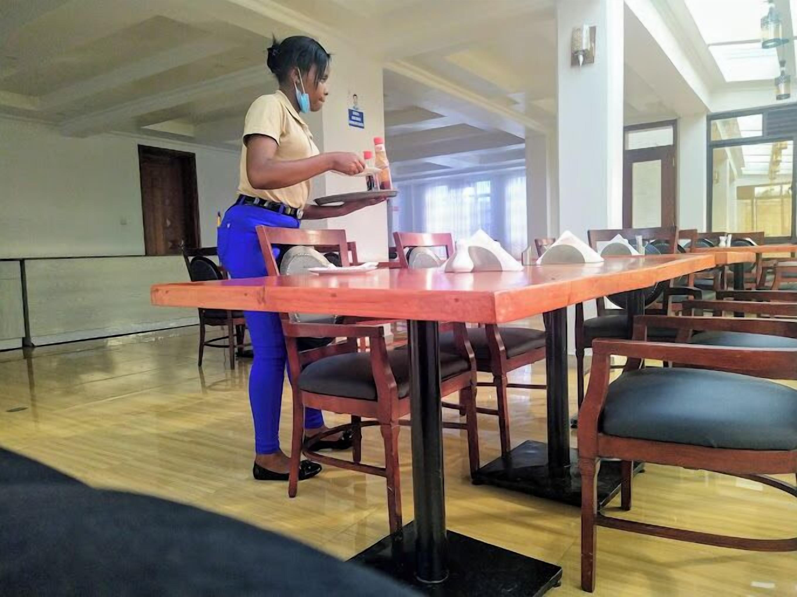 A spacious room with a long wooden table and chairs, a person serving food, and a large window providing natural light.
