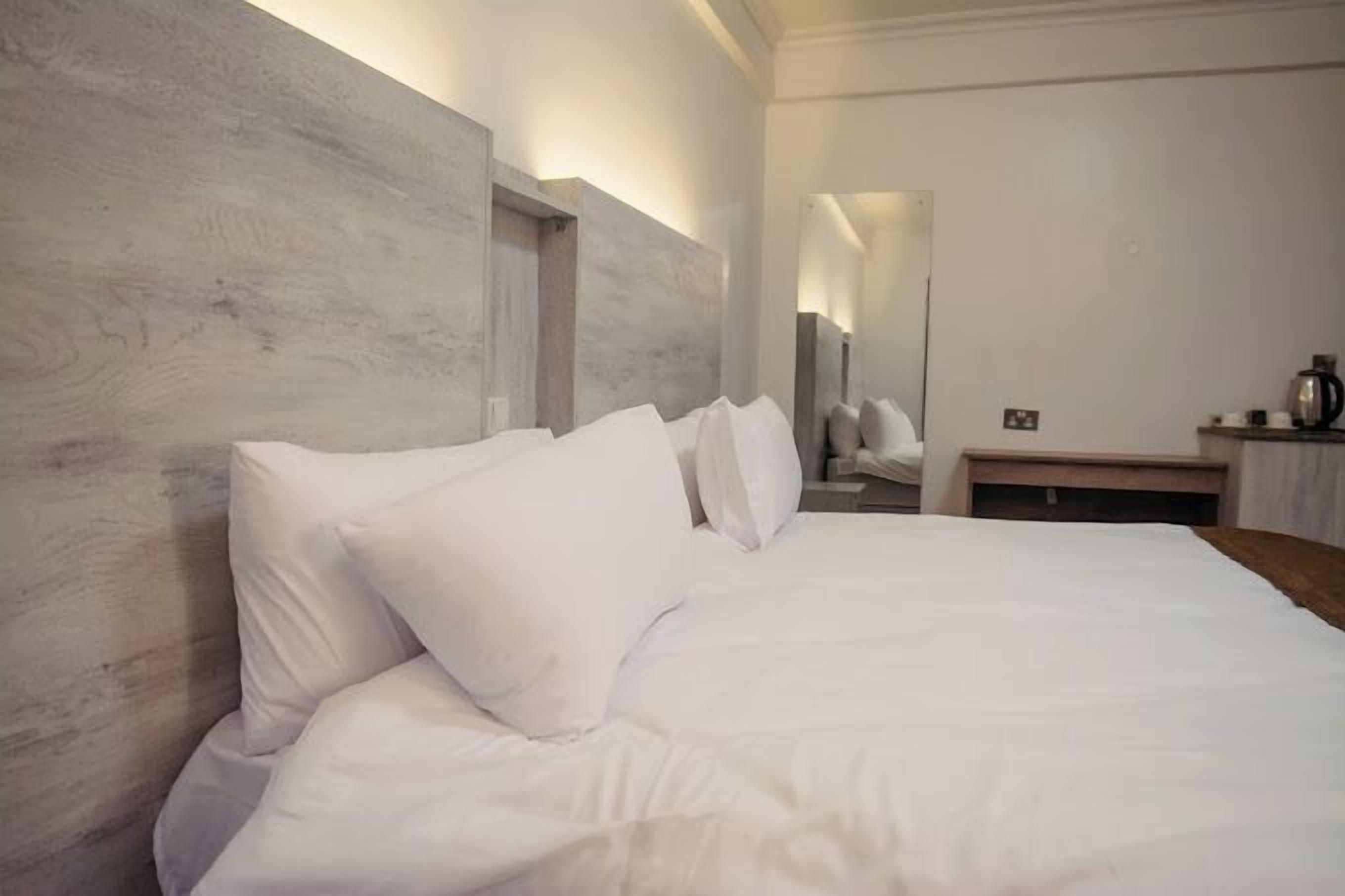 A hotel room with a large bed, a wooden headboard, and a small bedside table with a kettle.