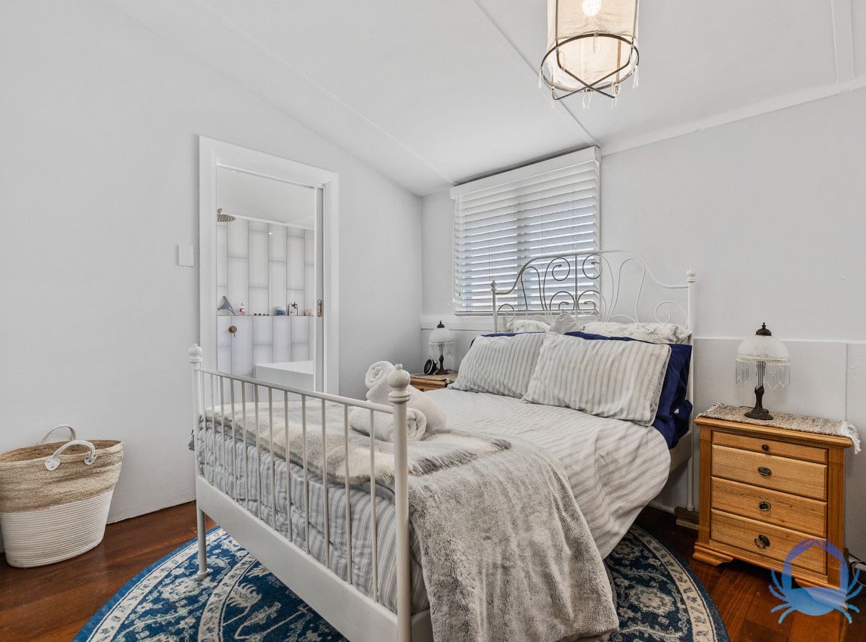 A bedroom with a white bed, a wooden nightstand, a lamp, a window with blinds, a radiator, and a blue patterned rug.