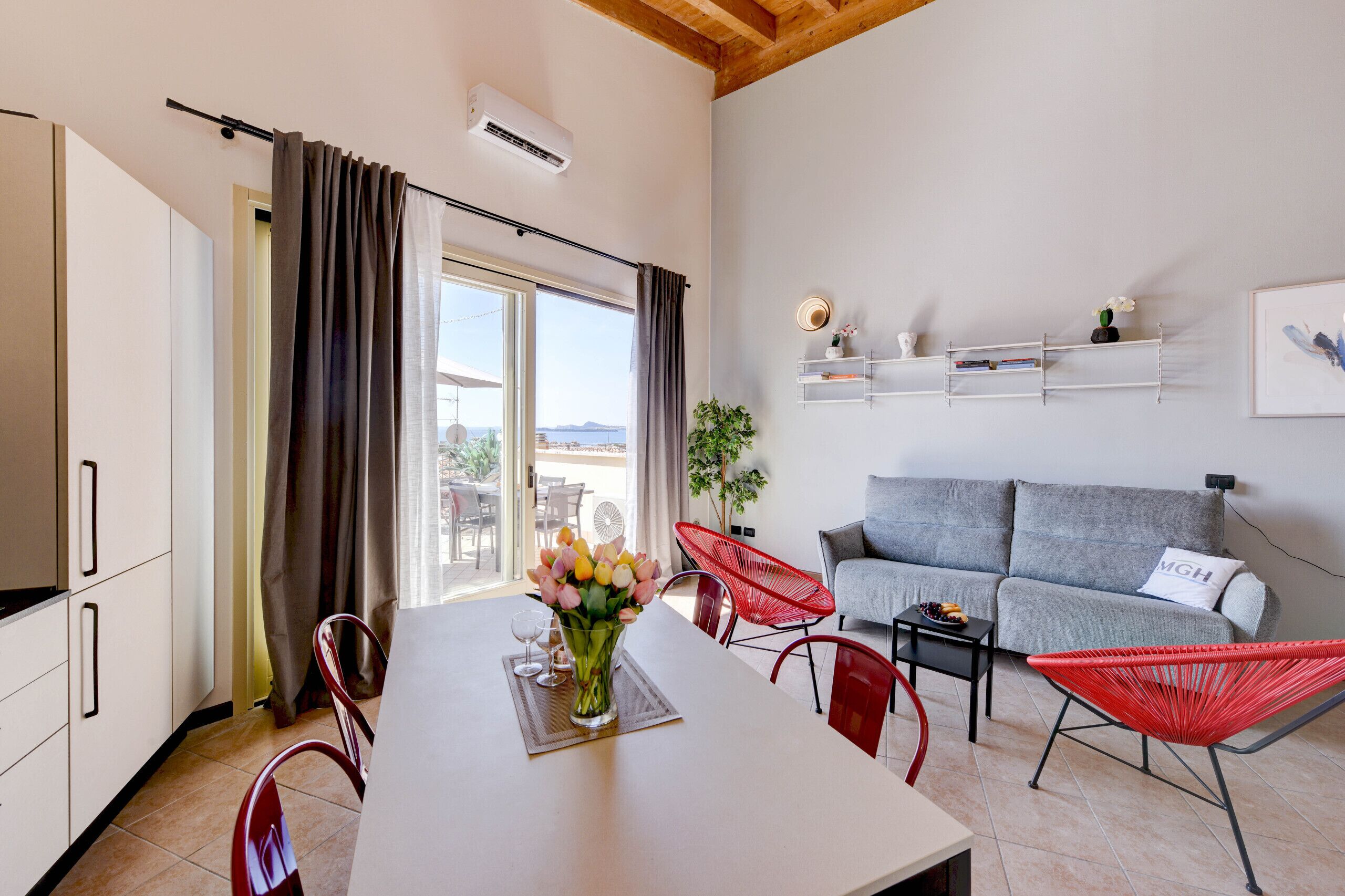 Un salon moderne avec un canapé, des fauteuils rouges, une table à manger ornée d’un vase de fleurs et une vue sur l’extérieur, visible à travers une porte vitrée.