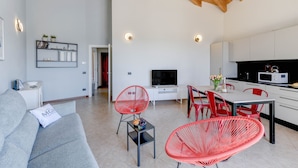 A modern living room with a grey sofa, red chairs, a TV, and a kitchen area with white cabinets and a microwave. - Palazzo Garda - Lakeview Penthouse 5 (Toscolano-Maderno)