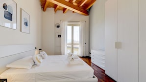 A bedroom with a bed, white walls, wooden beams, a white wardrobe, and artwork on the wall. - Palazzo Garda - Lakeview Penthouse 5 (Toscolano-Maderno)