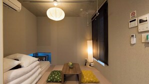 A room with tatami mats, a low wooden table, a bed with white bedding, a hanging light fixture, and a wall-mounted thermostat.