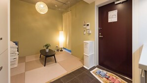A hotel room with tatami mats, a traditional Japanese-style lamp, a small table with a plant, a white refrigerator, and a dark wooden door.