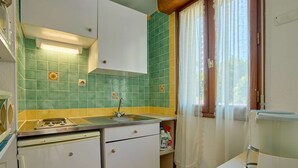 A compact kitchen with a sink, a stove, and a refrigerator. There is a window with curtains and a chair.