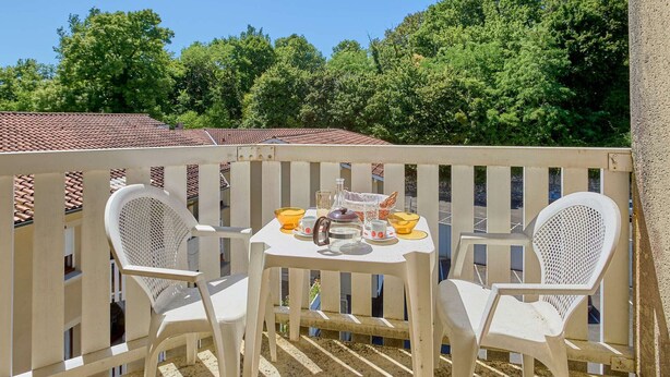 A balcony with a table set for two, white plastic chairs, and a view of a red-tiled roof and green trees.