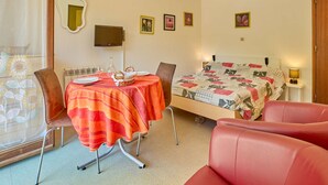 A bedroom with a bed, a table with a red tablecloth, two chairs, a television, and several framed pictures on the wall.