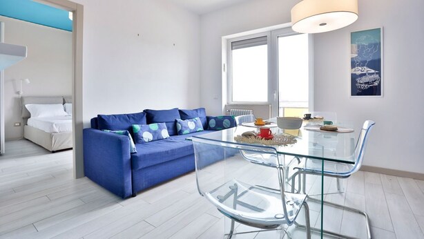 A modern living room with a blue sofa, a glass dining table, and a large window. - GuestHost - Vento di Mare House (Naples)