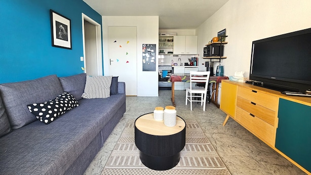 Una sala de estar con sofá, mesa de centro, televisor y una cocina al fondo.