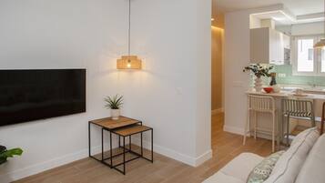 Een modern appartement met een flatscreen televisie aan de muur, een klein houten bijzettafeltje met een plant, een bank met gekleurde kussens, en een keuken die zichtbaar is door een open deur.