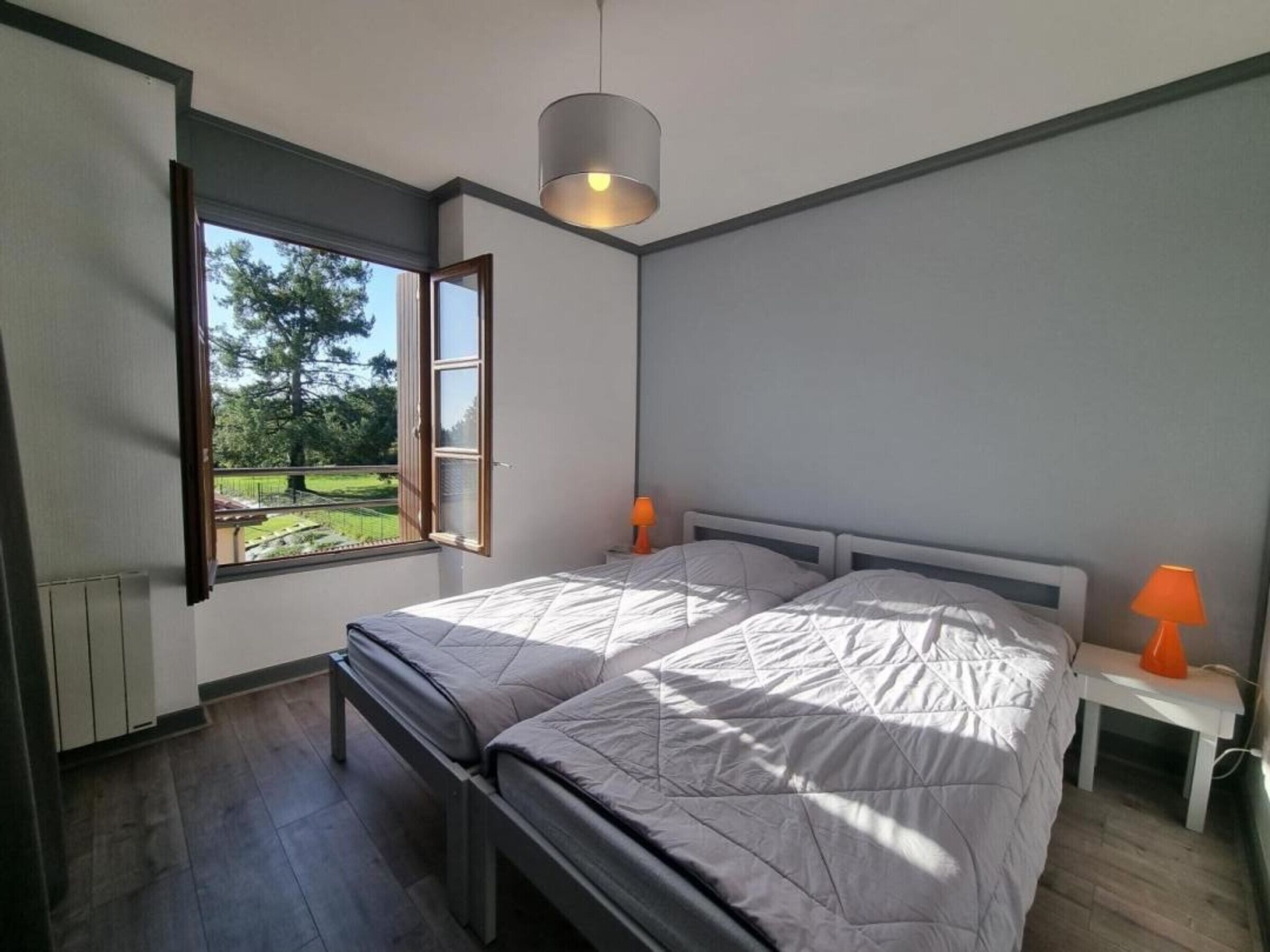 Ein modernes Schlafzimmer mit einem großen Bett, zwei Nachttischen mit Lampen, einem Fenster mit geöffnetem Klappladen und Blick auf einen Garten.