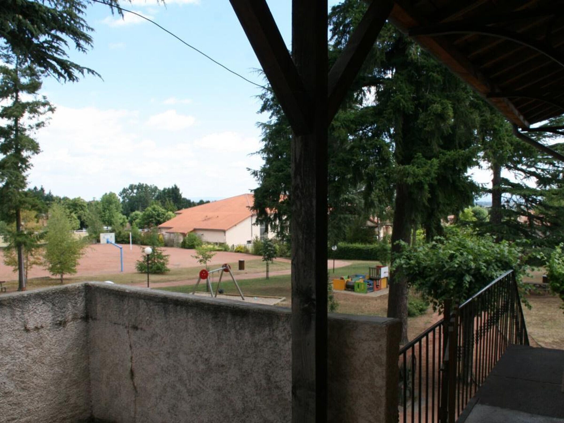 A covered outdoor area with a concrete wall, a metal railing, and a view of a building with a red roof and trees in the background.